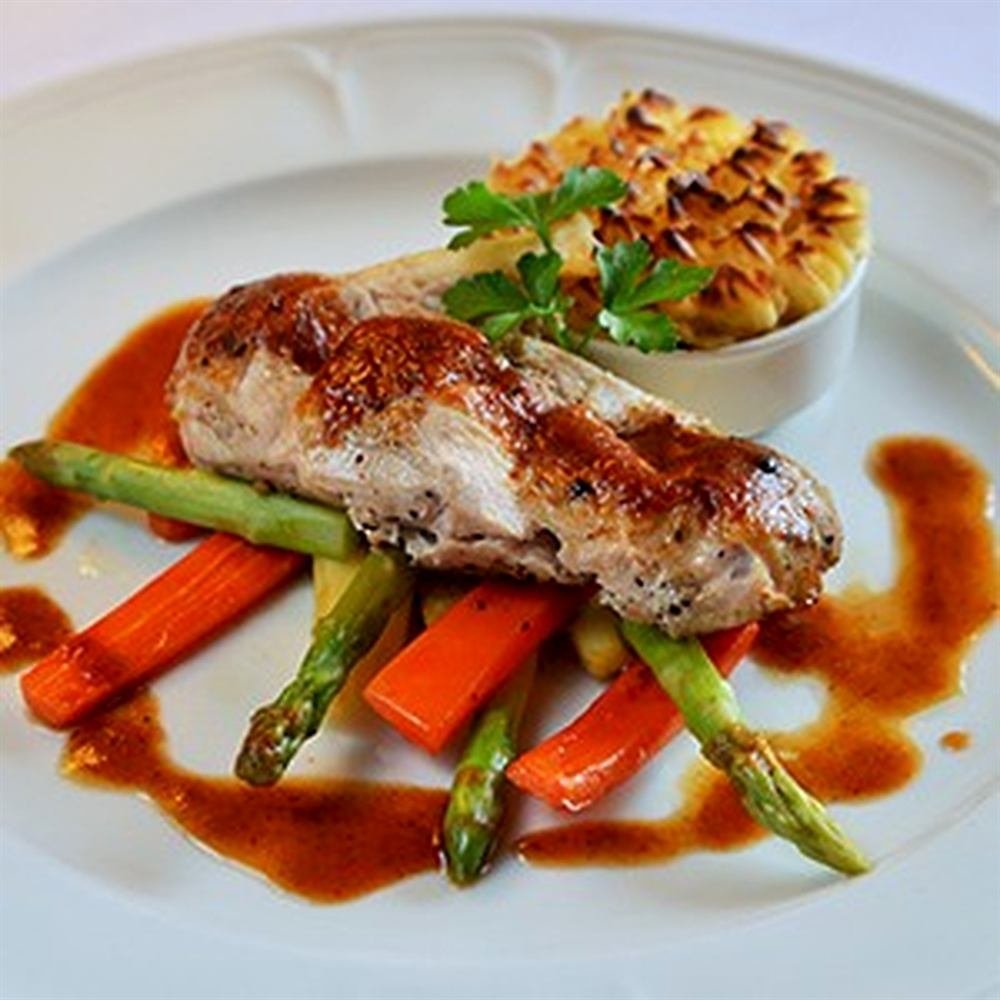 A plated main course of roast poultry with asparagus, carrots and a gratin, finished with a rich jus.